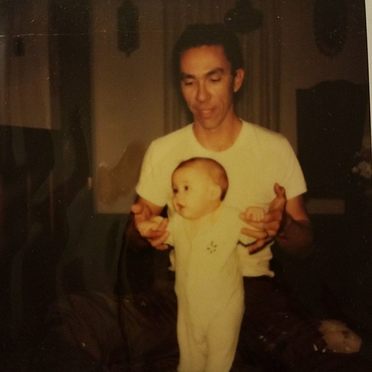 My father, John Almond, holding my hands as I stood as a baby. Picture is in a darkened living room. My father is wearing a white t-shirt and I am in a onesie.